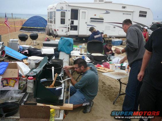People cooking at a beach campsite with an RV and tents