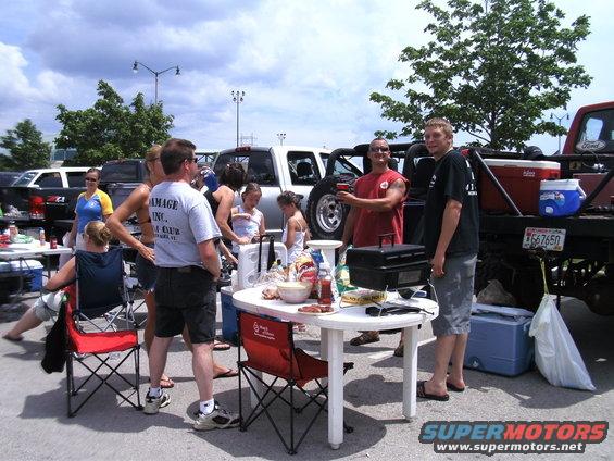 People gathered around a picnic table with food and grills, with several trucks visible in the background