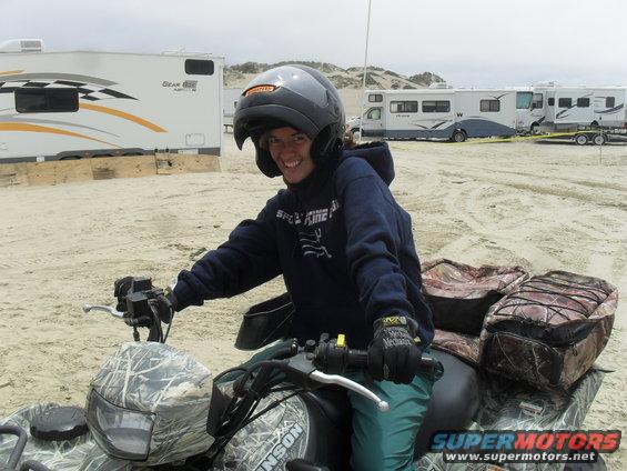Person wearing a helmet and hoodie on an ATV in a sandy area with RVs in the background