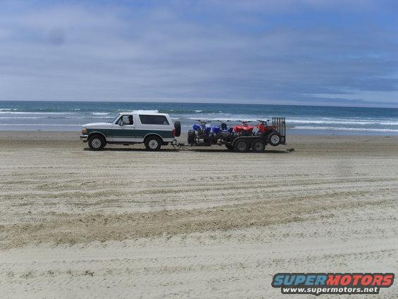 White and green Ford Bronco towing a trailer with ATVs on a sandy beach