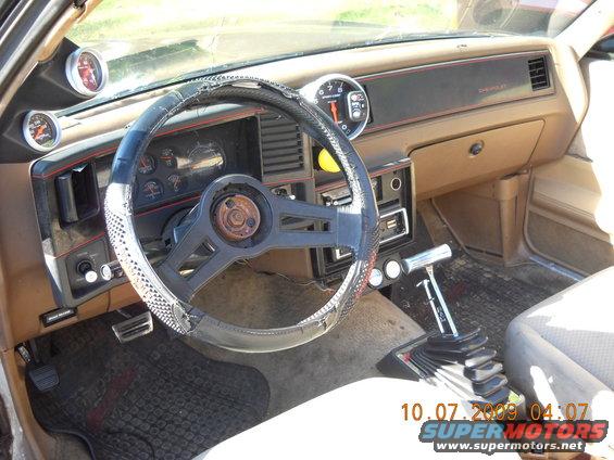 Car interior showing dashboard, steering wheel, aftermarket gauges, and floor shifter