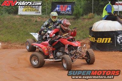 Two ATVs racing on a dirt track, one red and one white. Riders wear helmets and protective gear