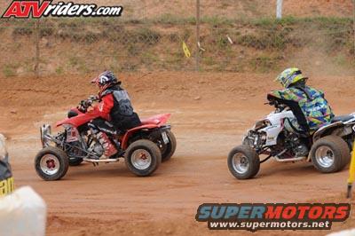 Two ATVs racing on a dirt track, one red and one white