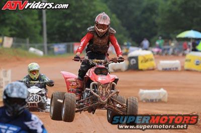 Rider on a red quad bike in a race, another rider on a white quad bike behind. Dirt track with barriers