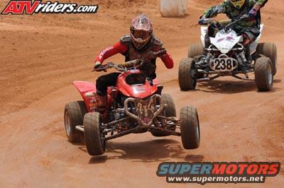 Red ATV with rider in red gear in foreground, white ATV with rider in green and black gear in background on dirt track
