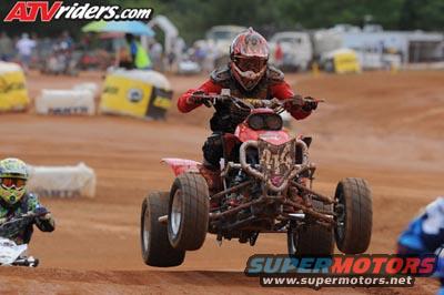 ATV rider in red gear jumps over a dirt track, another rider visible to the left