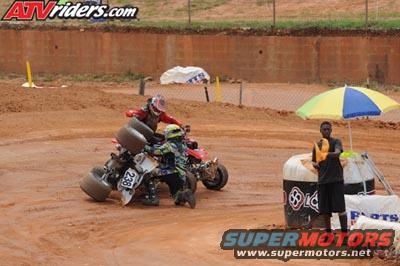Two ATVs on a dirt track, one overturned with a rider on the ground, the other upright with a rider