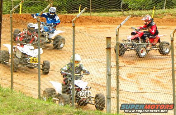 Four ATVs racing on a dirt track, seen through a fence. One ATV has number 238, another has 44