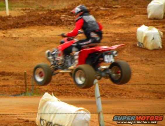 Red ATV with rider in helmet and red jacket mid-air on a dirt track. Number 414 visible on rear fender