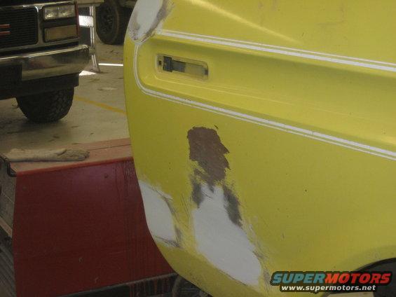 Yellow vehicle fender with body filler and primer, showing damage and repair work. A GMC truck is partially visible to the left