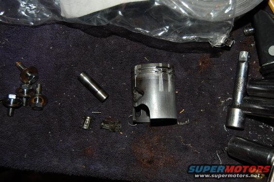 Damaged piston and assorted bolts and tools on a dark cloth surface
