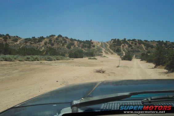 Dark vehicle hood in foreground, dirt road leading to desert hills under blue sky