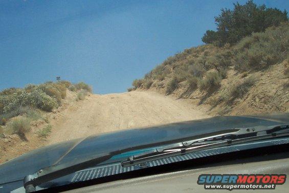 Dark dashboard and windshield wipers on a sunny day, looking up a dirt road on a dry, brush-covered hill