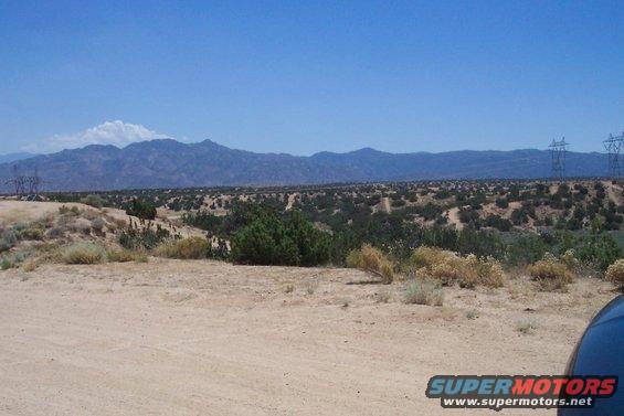 Desert landscape with mountains, scrub brush, and power lines under a clear blue sky. Part of a dark vehicle is visible on the right