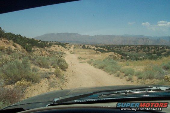 View from inside a vehicle looking out at a dirt road winding through a desert landscape with scrub brush and distant mountains under a clear blue sky