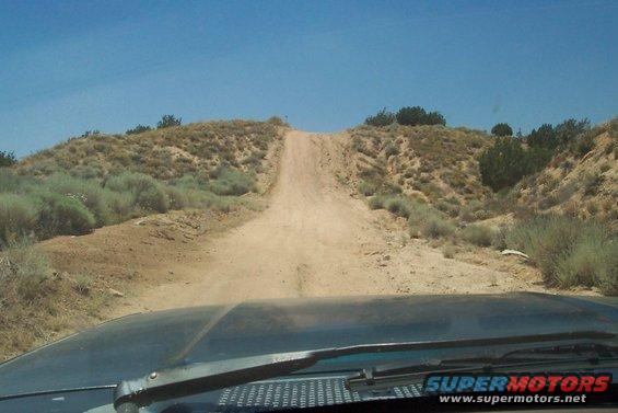 View from inside a vehicle looking up a dusty dirt road on a hill under a clear blue sky