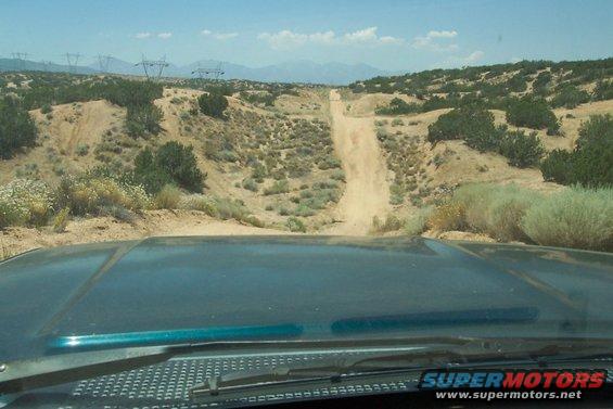 View from inside a vehicle looking out at a dusty, uphill dirt road in a desert landscape with power lines in the distance