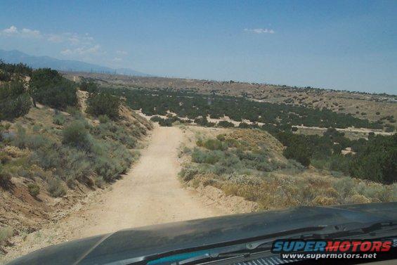 Dirt road winds through a desert landscape with sparse vegetation and distant mountains under a blue sky. Supermotors.net logo visible