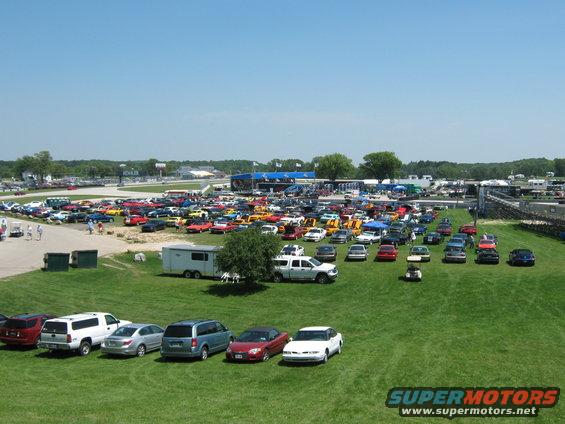 many cars parked on grass at a raceway with buildings and track in background