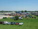 1985 Ford Mustang - A large gathering of vehicles are parked on a grassy area next to a race track on a sunny day.