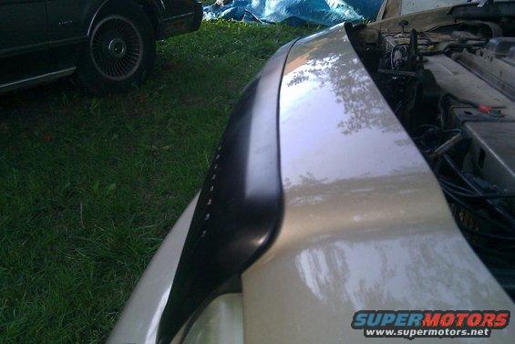 close-up of car fender and open hood showing engine components, another car visible in background