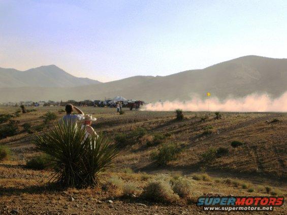 Spectators watch a dusty desert race with multiple vehicles in the distance