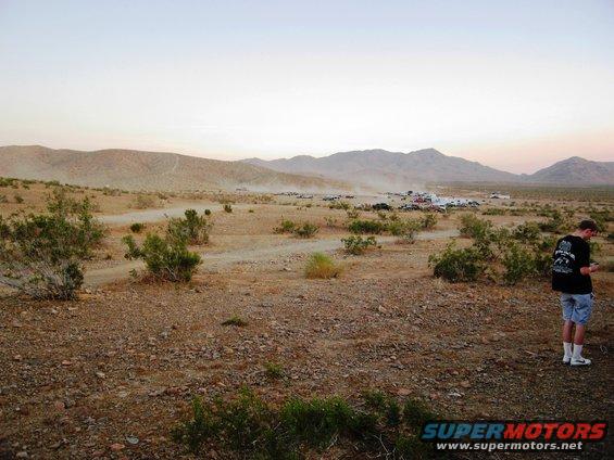 Desert scene with distant vehicles and a man in the foreground, with 'SUPERMOTORS' text at the bottom right