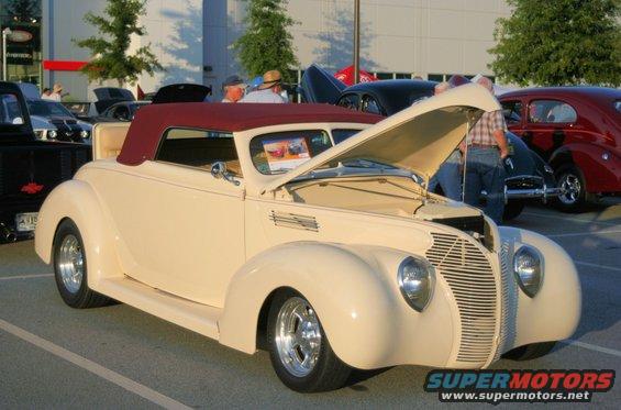 Cream convertible with maroon top, hood open, parked at car show. Other classic cars visible