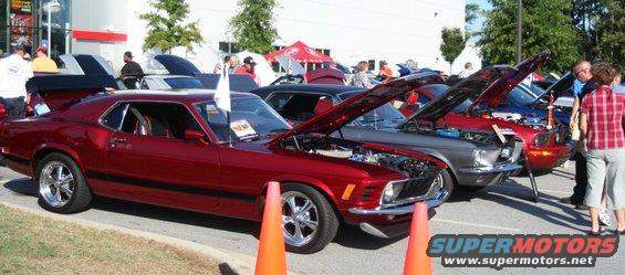 Red classic car with hood open, parked in a line with other cars. Orange cones in foreground