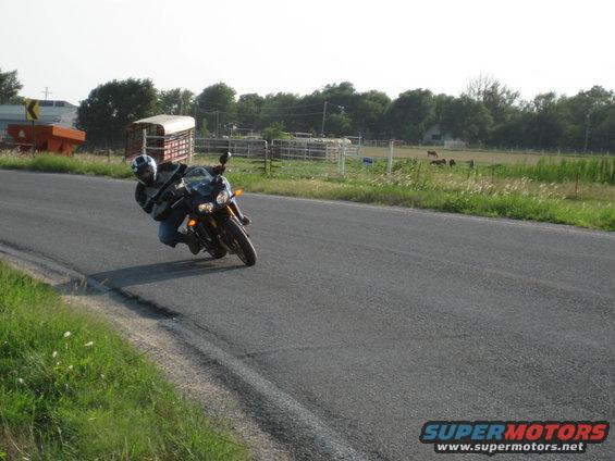 Motorcycle leaning into a turn on a paved road, with farm fields and trees in the background