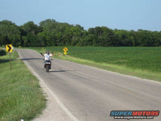 Motorcycle rider on a road with arms raised, surrounded by fields and trees