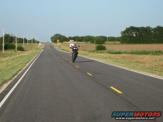 Motorcycle on one wheel on a highway, rider visible, blue sky, fields on the side
