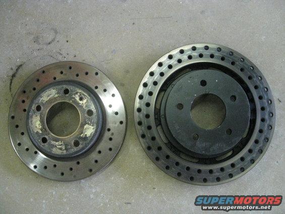 Two vented brake rotors, one with drilled holes and a worn hub, the other with drilled holes and a black hat, on a concrete surface