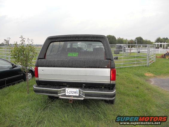 Rear view of a black and silver Ford Bronco with a camper shell, parked on grass near a metal fence