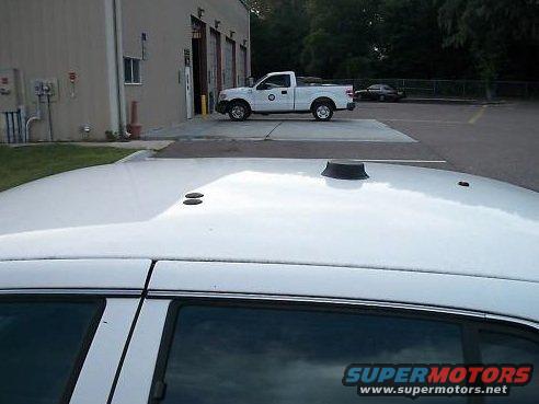 Top view of a white sedan's roof with an antenna; a white pickup truck is parked in front of a building in the background