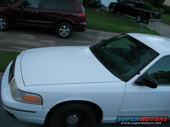White sedan, possibly a Ford Crown Victoria, parked on a driveway. A maroon SUV and a black pickup truck are also visible