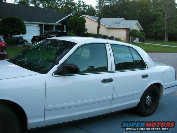 White sedan parked in driveway, side view. Houses and trees in background