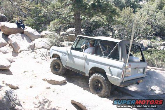 Gray Ford Bronco driving on a rocky trail. A person sits on a rock nearby