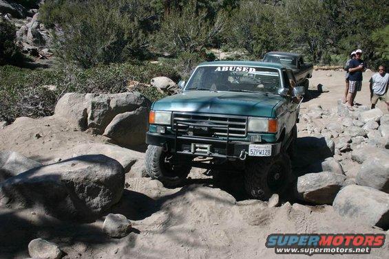 Green Ford F-Series truck off-roading over rocks. Another truck is behind it. People are watching