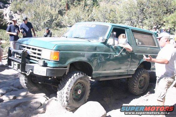 Green Ford Bronco with off-road tires on a rocky trail. People are standing around it