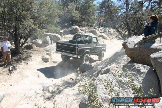 Ford Bronco driving on a rocky trail, kicking up dust. Spare tire mounted in the bed