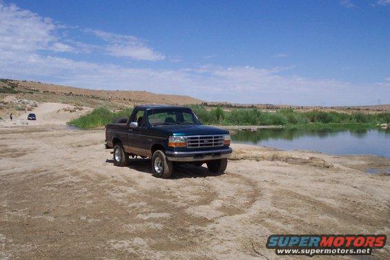 Dark blue Ford F-150 pickup truck parked on dirt near a body of water. Another vehicle is visible in the background