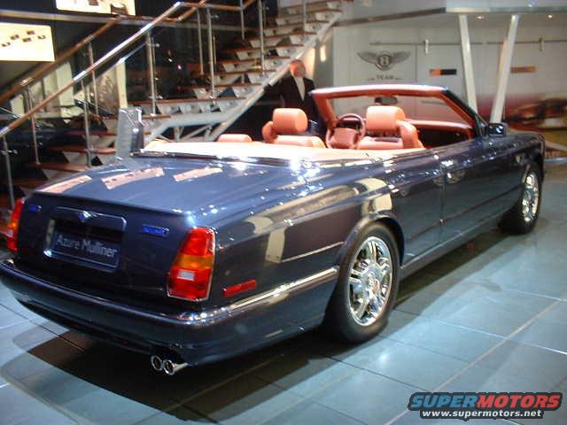 Dark blue Bentley Azure Mulliner convertible with tan interior, rear 3/4 view, chrome wheels, dual exhaust
