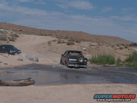 Rear view of a 1995 Ford Bronco driving through water, with a black sedan on the left