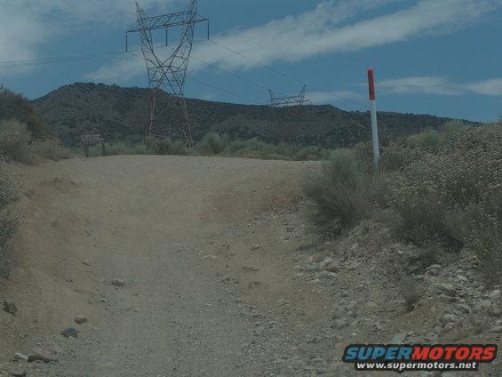 Dirt road leading uphill toward mountains with power lines and towers. A red and white pole is on the right