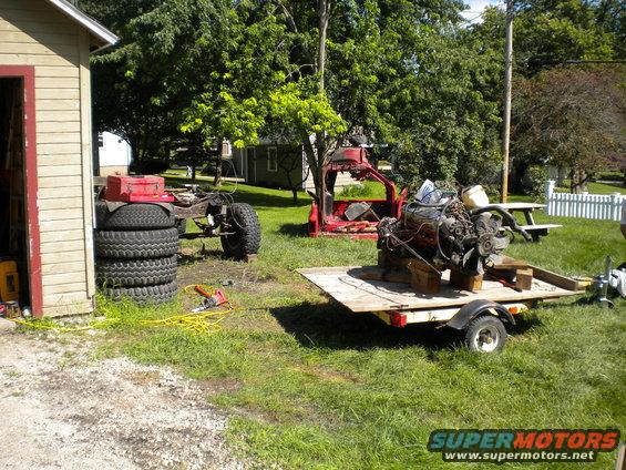 Disassembled red pickup truck chassis with stacked tires and engine on trailer