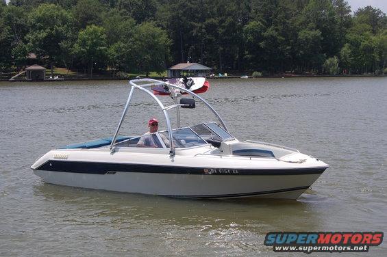 White boat with blue interior trim and a metal frame with a wakeboard on top, moving on a lake