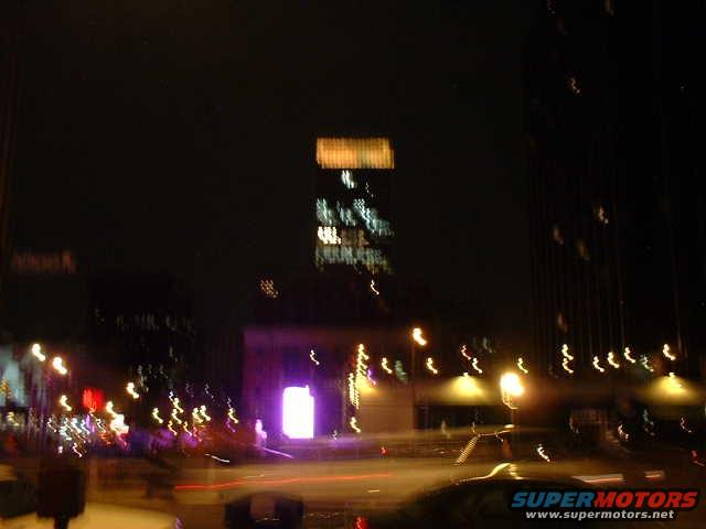 Blurry dark scene with streetlights and building lights, a dark car is partially visible in the foreground