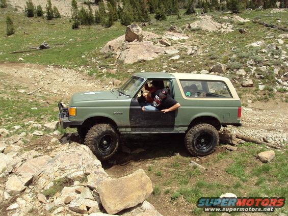 Green Ford Bronco with a tan top navigating a rocky mountain trail, driver leaning out the window