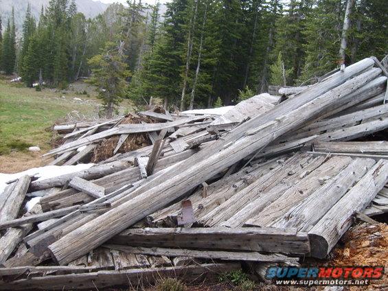 pile of weathered lumber and debris in a wooded area with snow patches